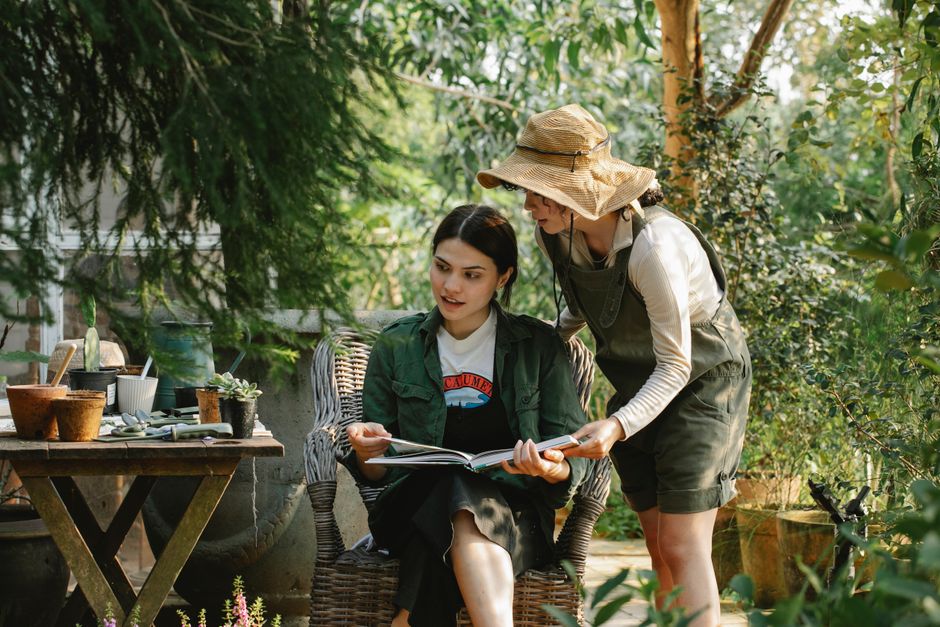Two women in an indoor garden shop are engaged in garden maintenance activities amidst a lush display of various plants, including large leafy tropical species and smaller potted plants. The woman on 